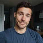 A smiling young man with short brown hair wearing a blue shirt, indoors with soft lighting.