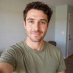 Smiling man in a casual green t-shirt taking a selfie indoors with natural lighting.